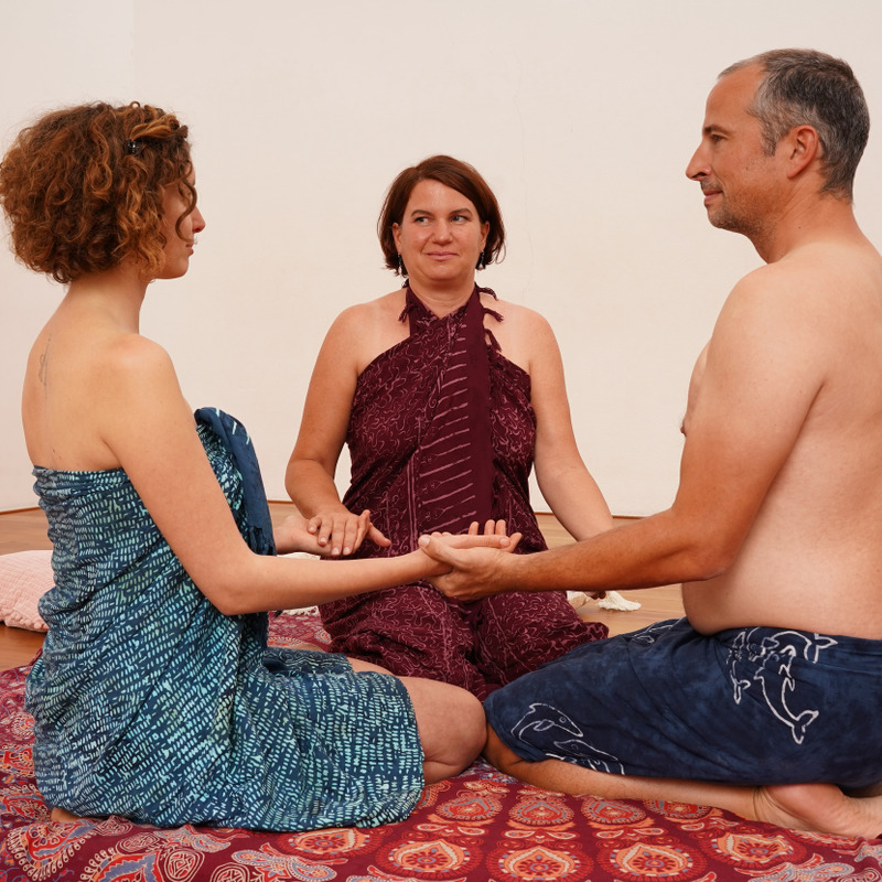 Zwei Frauen und ein Mann sitzen am Boden, halten sich an den Händen, nur mit Lunghis bekleidet