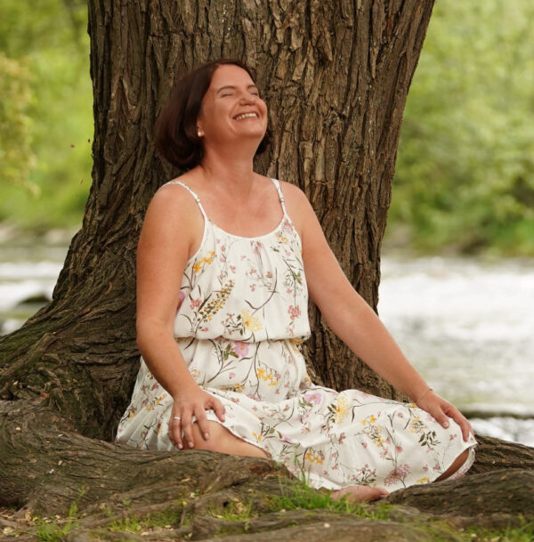 Frau sitzt im Schneidersitz vor einem Baum, lacht