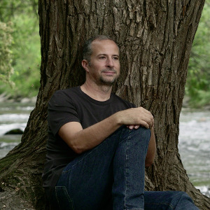 Mann sitzt vor einem Baum, eine Hand auf dem angewinkelten Knie abgelegt